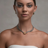 A woman with her hair pulled back wears a delicate silver chain necklace with a gold-toned textured center bead. She is wearing a white strapless top against a neutral gray studio background.
