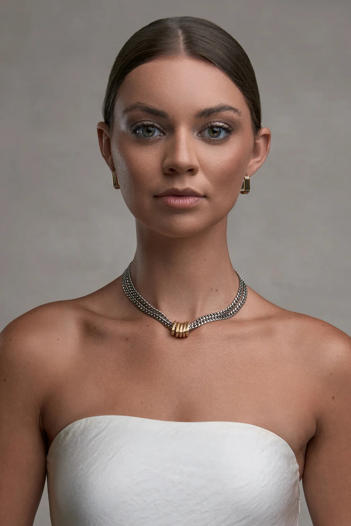 A woman with her hair pulled back wears a delicate silver chain necklace with a gold-toned textured center bead. She is wearing a white strapless top against a neutral gray studio background.