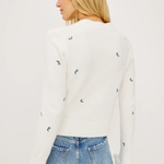 A woman shown from the back wearing a white long-sleeve knit sweater with small scattered blue and yellow embroidered details, paired with light blue jeans, against a plain white studio background.