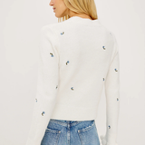 A woman shown from the back wearing a white long-sleeve knit sweater with small scattered blue and yellow embroidered details, paired with light blue jeans, against a plain white studio background.