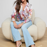 Woman sitting on a beige armchair wearing a colorful blouse and light blue jeans.