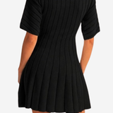 A woman wearing a black short-sleeve ribbed pleated mini dress, shown from the back against a light gray studio background.