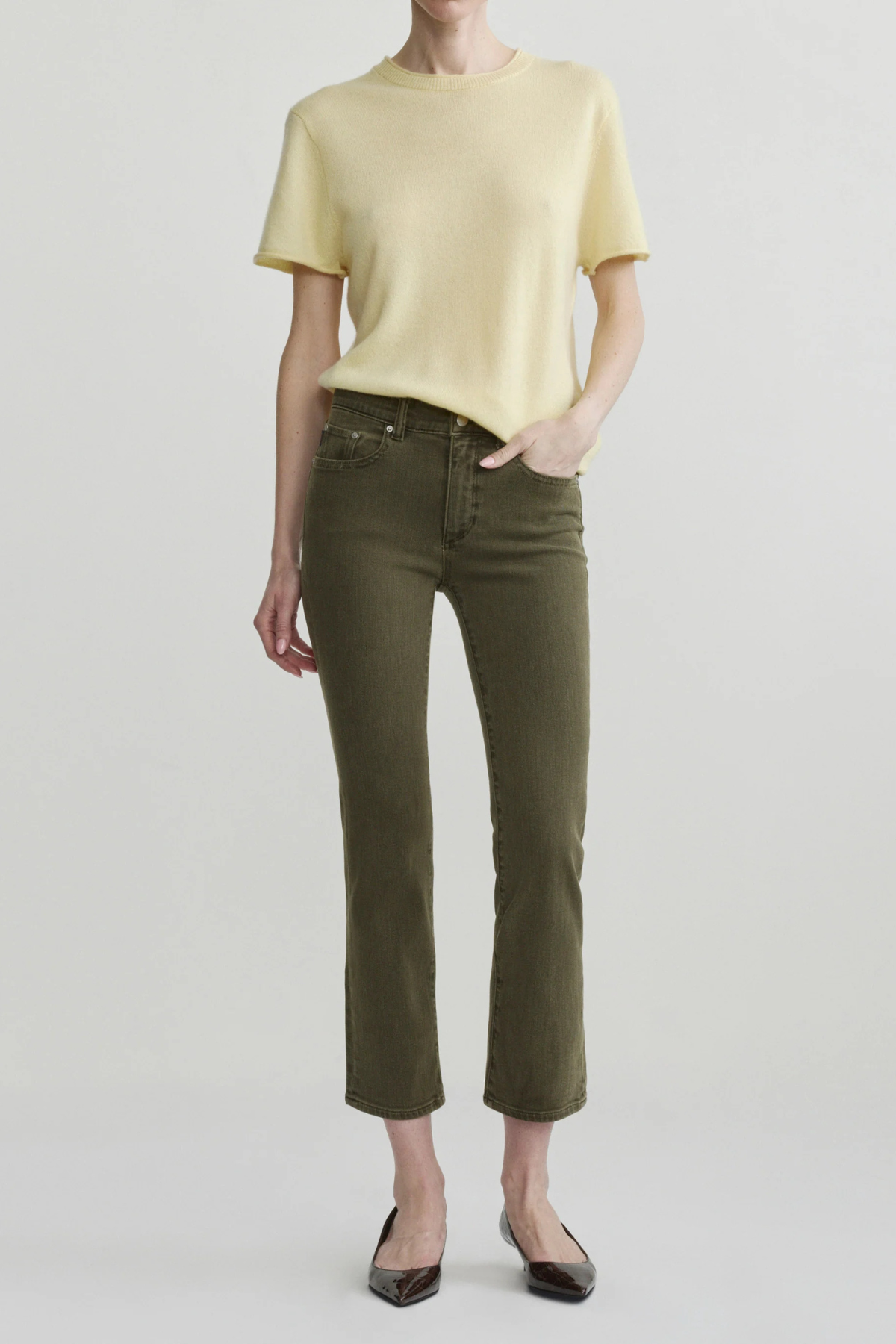 A model wearing a pale yellow short-sleeve knit top and olive green cropped jeans with dark brown pointed flats, standing against a light gray studio background.