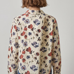 A person shown from the back wearing a cream-colored long-sleeve button-up shirt with a red and purple floral print, paired with blue jeans, against a plain light gray background.