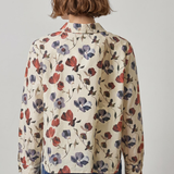 A person shown from the back wearing a cream-colored long-sleeve button-up shirt with a red and purple floral print, paired with blue jeans, against a plain light gray background.