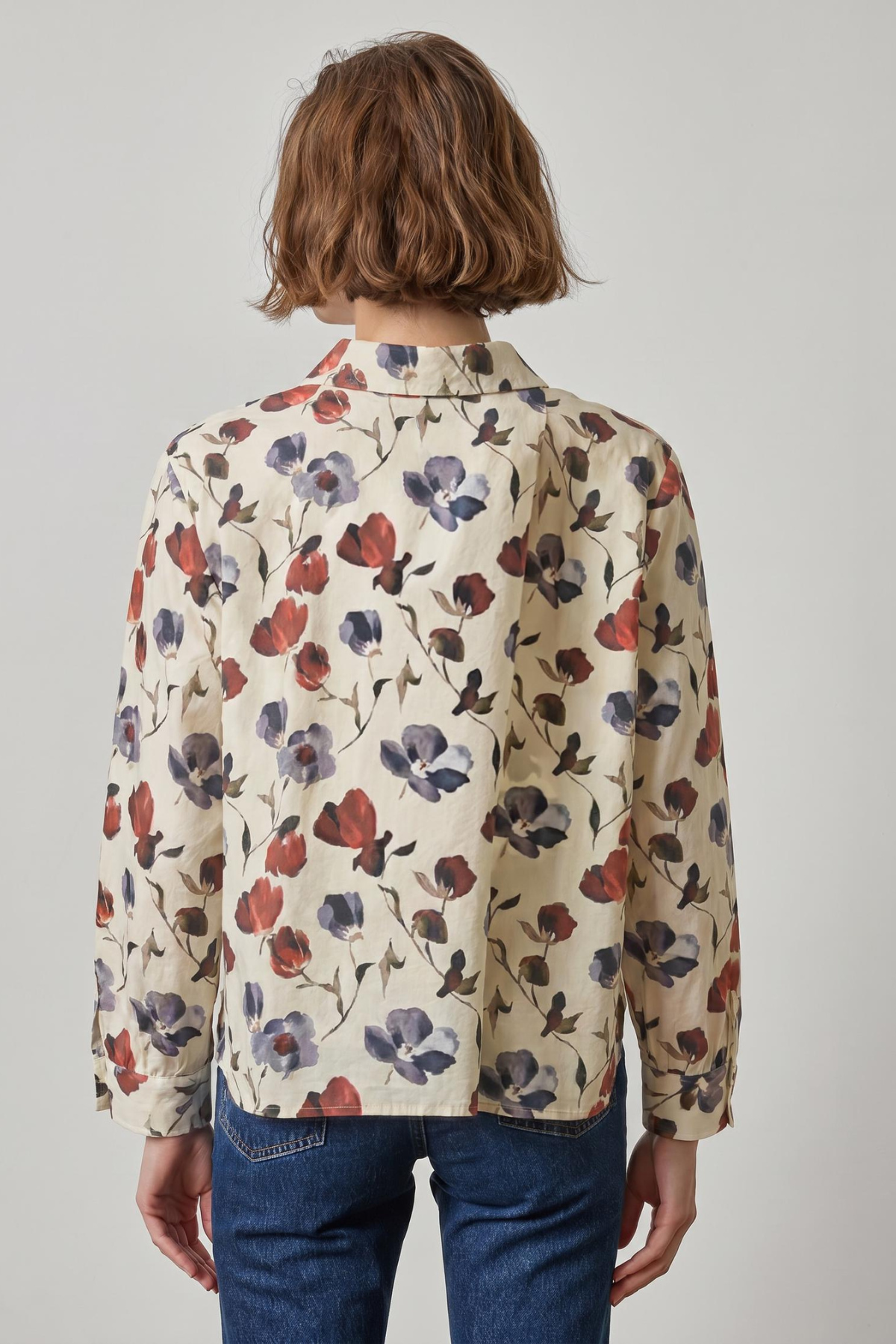 A person shown from the back wearing a cream-colored long-sleeve button-up shirt with a red and purple floral print, paired with blue jeans, against a plain light gray background.