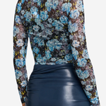 A woman shown from the back wearing a sheer long-sleeve floral mesh top with blue, purple, and black tones, paired with a dark navy faux-leather skirt, against a light gray studio background.