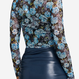A woman shown from the back wearing a sheer long-sleeve floral mesh top with blue, purple, and black tones, paired with a dark navy faux-leather skirt, against a light gray studio background.
