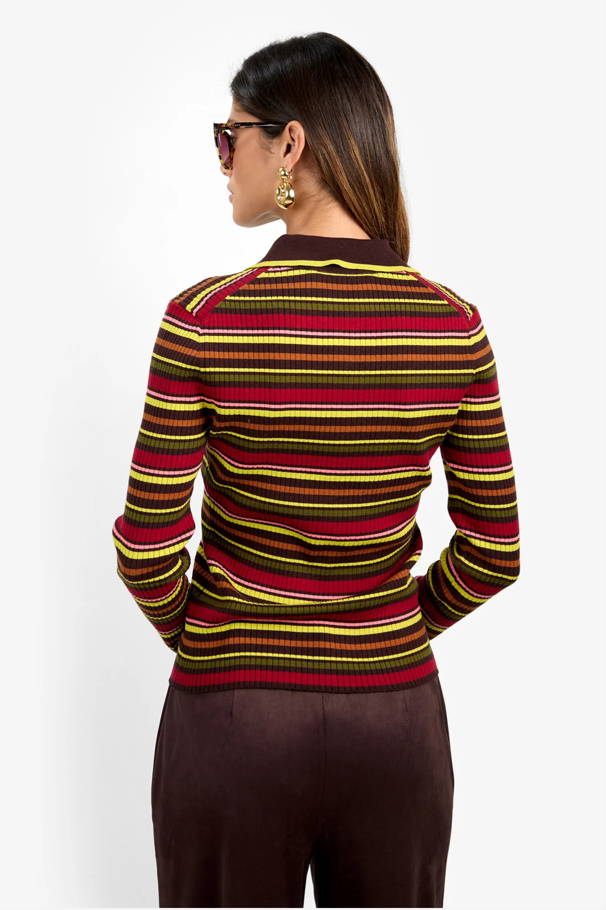 A woman shown from the back wearing a fitted long-sleeve knit sweater with horizontal stripes in red, yellow, and brown over a dark base, paired with dark brown pants against a plain light background.