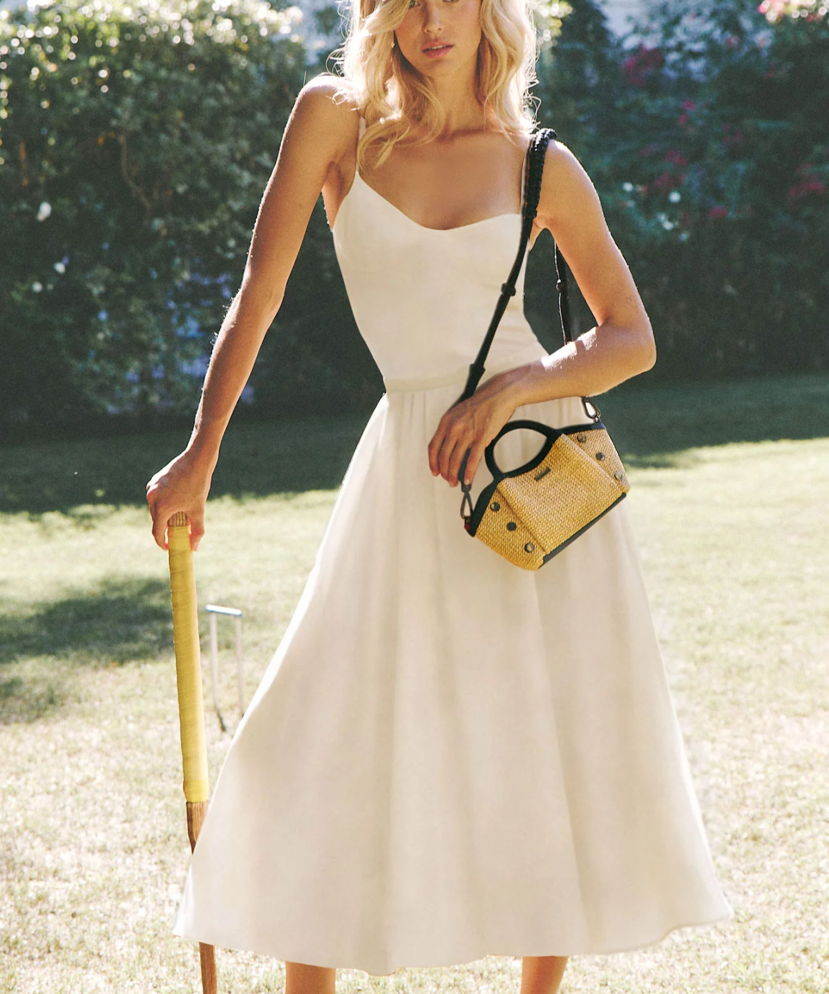 Woman in a white dress holding a yellow bag and a cane on a grassy lawn.