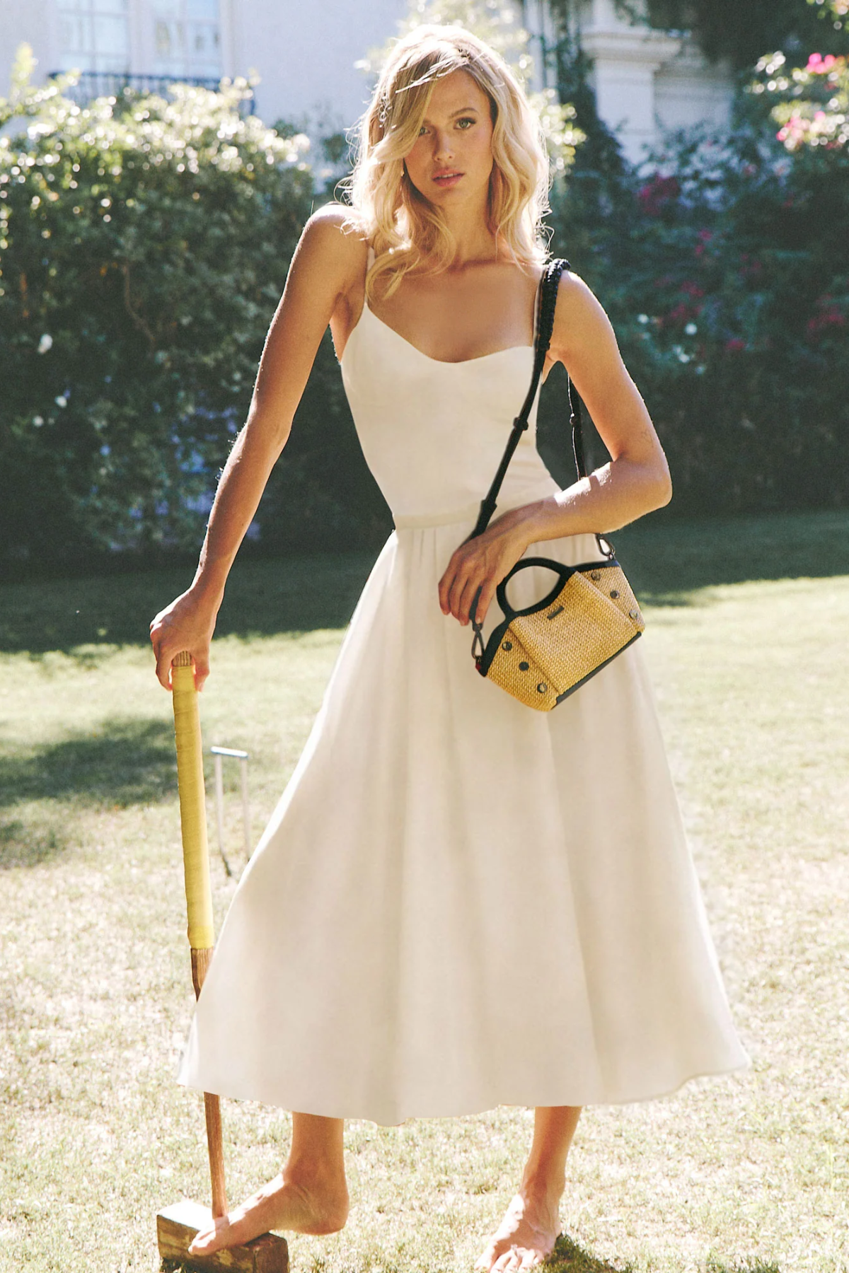 Woman in a white dress holding a yellow bag and a cane on a grassy lawn.