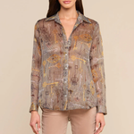 A woman modeling a long-sleeve button-up shirt with a brown, beige, and gold geometric pattern, paired with beige pants, against a solid beige background.