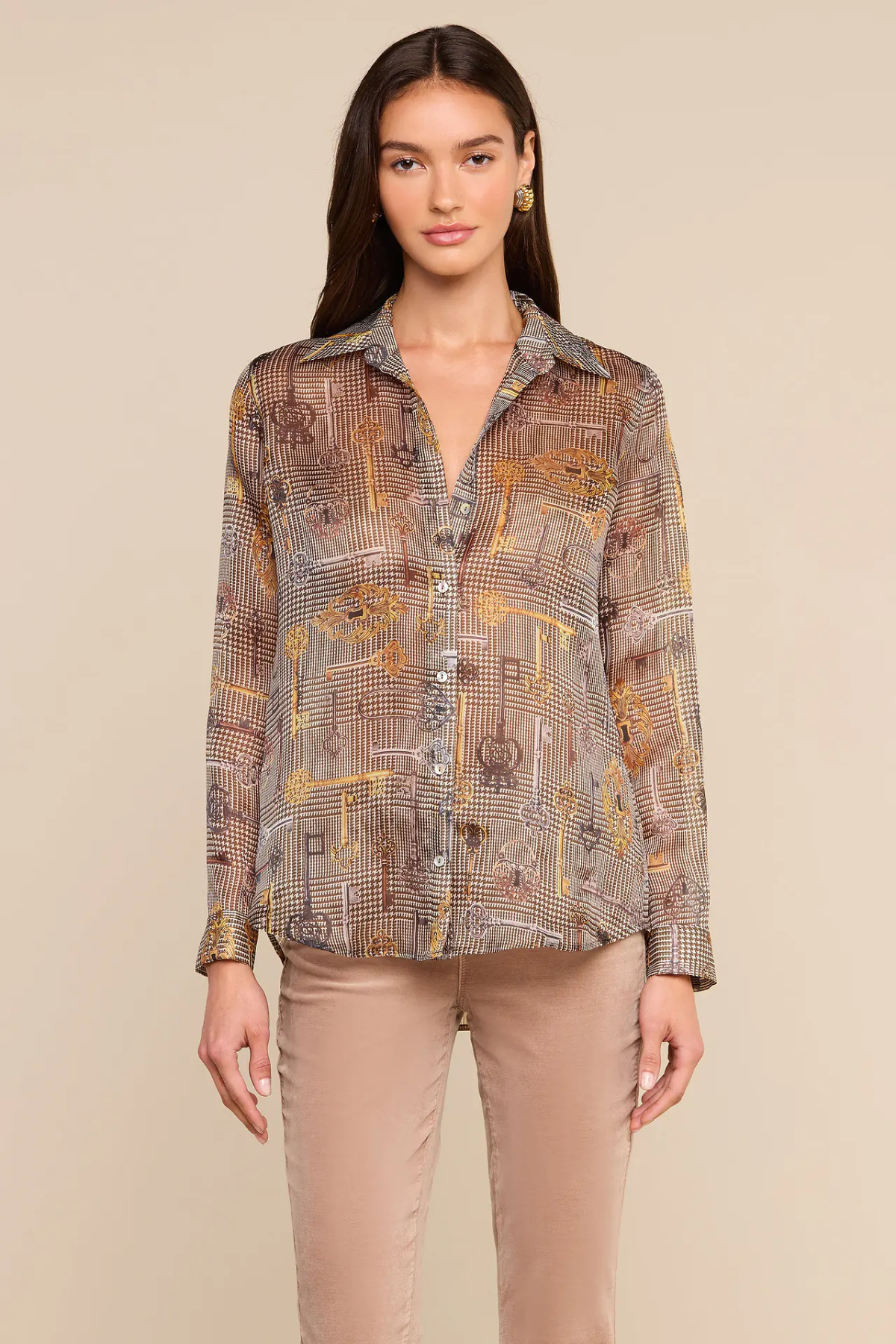 A woman modeling a long-sleeve button-up shirt with a brown, beige, and gold geometric pattern, paired with beige pants, against a solid beige background.