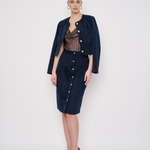 A model stands against a light gray studio background wearing a dark navy blue two-piece set consisting of a cropped button-front jacket and a matching knee-length skirt, layered over a patterned top and styled with nude high heels.