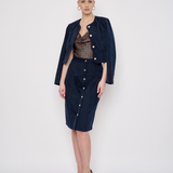 A model stands against a light gray studio background wearing a dark navy blue two-piece set consisting of a cropped button-front jacket and a matching knee-length skirt, layered over a patterned top and styled with nude high heels.