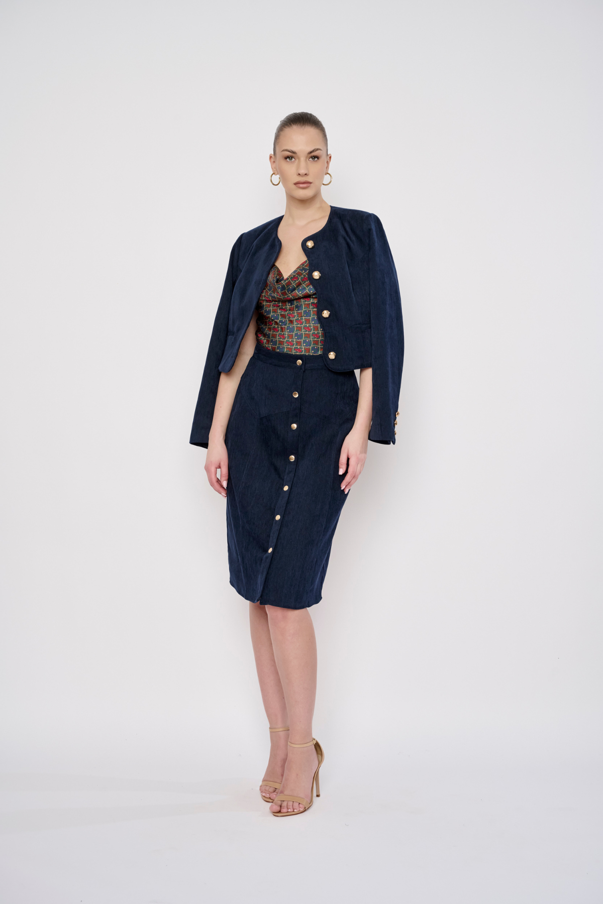 A model stands against a light gray studio background wearing a dark navy blue two-piece set consisting of a cropped button-front jacket and a matching knee-length skirt, layered over a patterned top and styled with nude high heels.