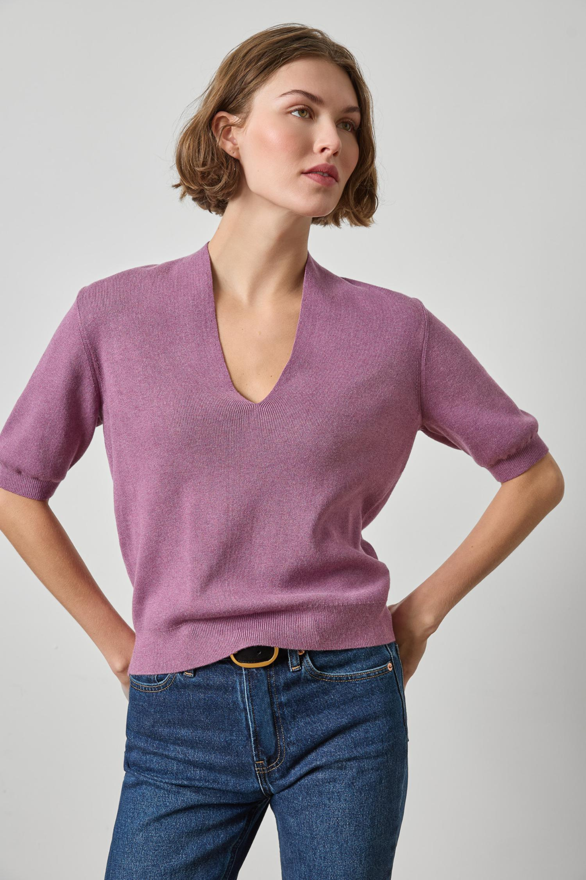 A woman modeling a short-sleeve V-neck knit sweater in dusty mauve purple, paired with blue jeans, against a light gray studio background.