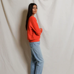 Person wearing a red sweater and blue jeans standing against a white draped curtain.