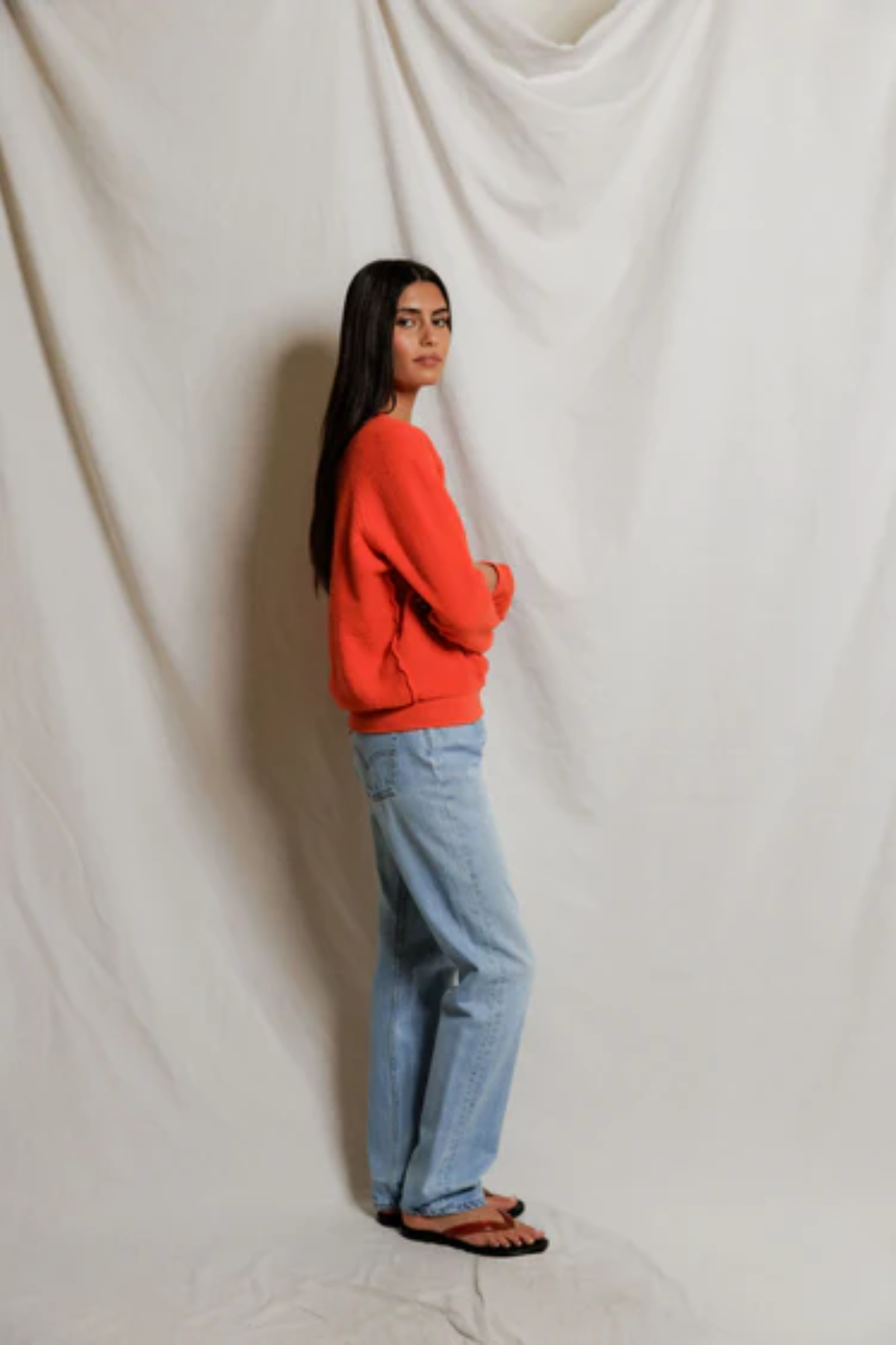Person wearing a red sweater and blue jeans standing against a white draped curtain.