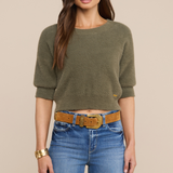 Model wearing an olive green sweater with jeans and a belt on a beige background.