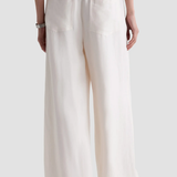 A person shown from the back wearing loose, wide-leg pants in a warm off-white or cream color, with an elastic waistband and two large patch pockets, against a light gray studio background.