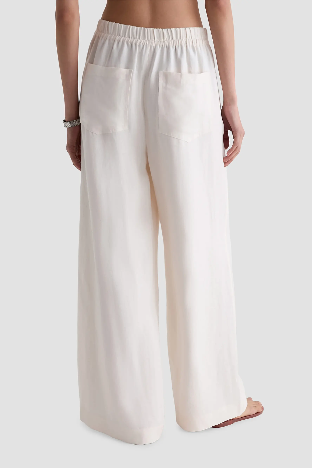 A person shown from the back wearing loose, wide-leg pants in a warm off-white or cream color, with an elastic waistband and two large patch pockets, against a light gray studio background.