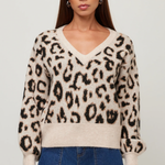 A woman modeling a fuzzy knit V-neck sweater with a beige, brown, and black leopard print pattern, paired with blue jeans, against a light gray studio background.