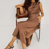 A woman with long curly dark hair sits on a brown leather chair wearing a sleeveless, high-neck brown satin midi dress and black strappy high-heeled sandals against a light gray studio background.