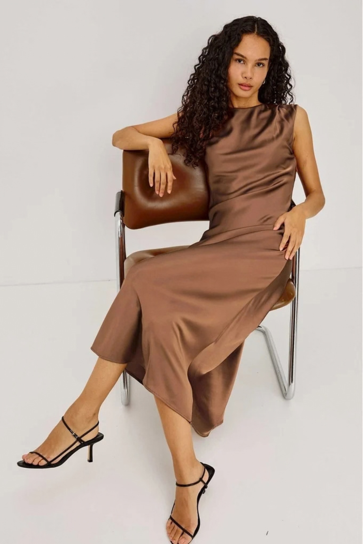 A woman with long curly dark hair sits on a brown leather chair wearing a sleeveless, high-neck brown satin midi dress and black strappy high-heeled sandals against a light gray studio background.
