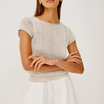 Woman wearing a beige knit top and white shorts with sunglasses on a plain background