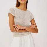 Woman wearing a beige knit top and white shorts with sunglasses on a plain background