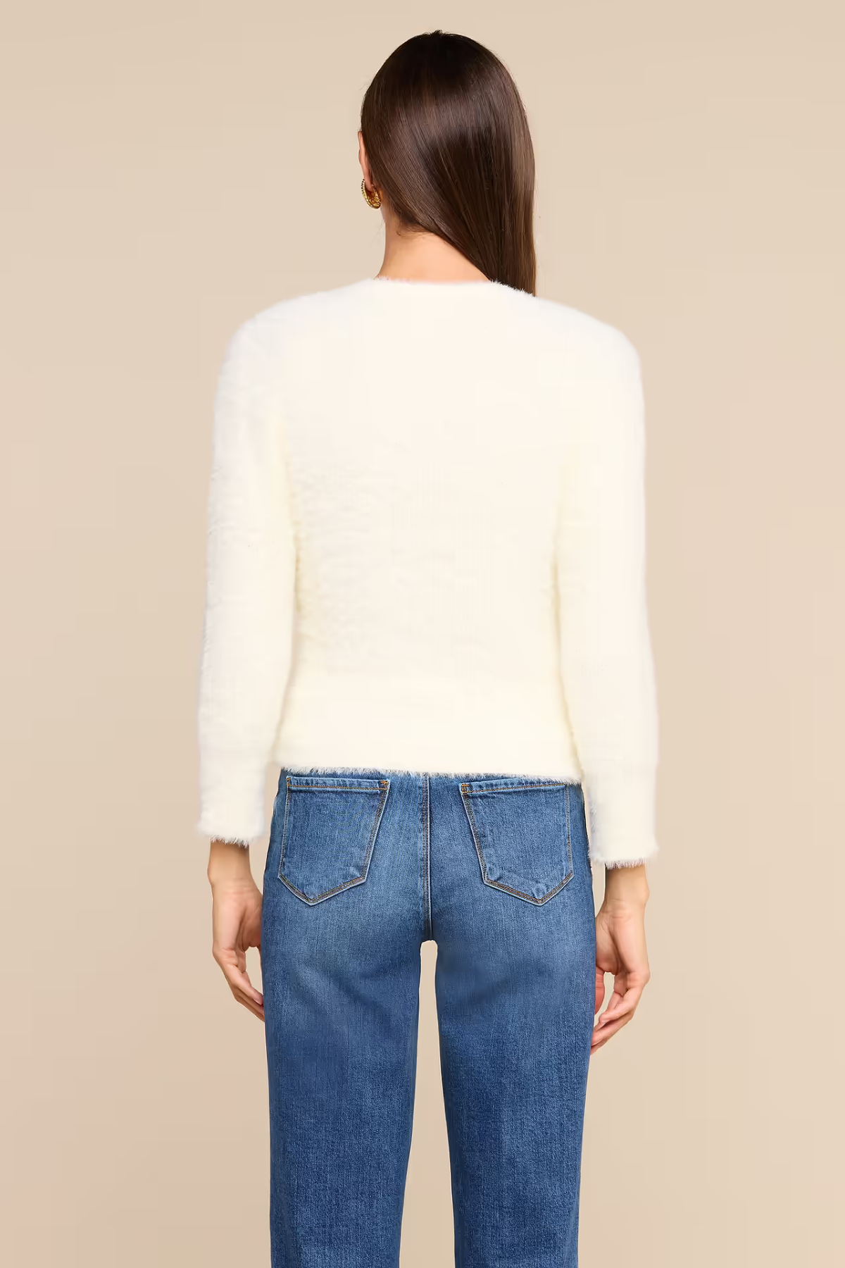 A woman shown from the back wearing a cream-colored, long-sleeve fuzzy knit top and blue jeans against a solid beige background.