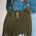 An olive green suede fringe handbag with gold-tone hardware, held by a person wearing a blue denim jacket and jeans.