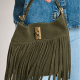 An olive green suede fringe handbag with gold-tone hardware, held by a person wearing a blue denim jacket and jeans.