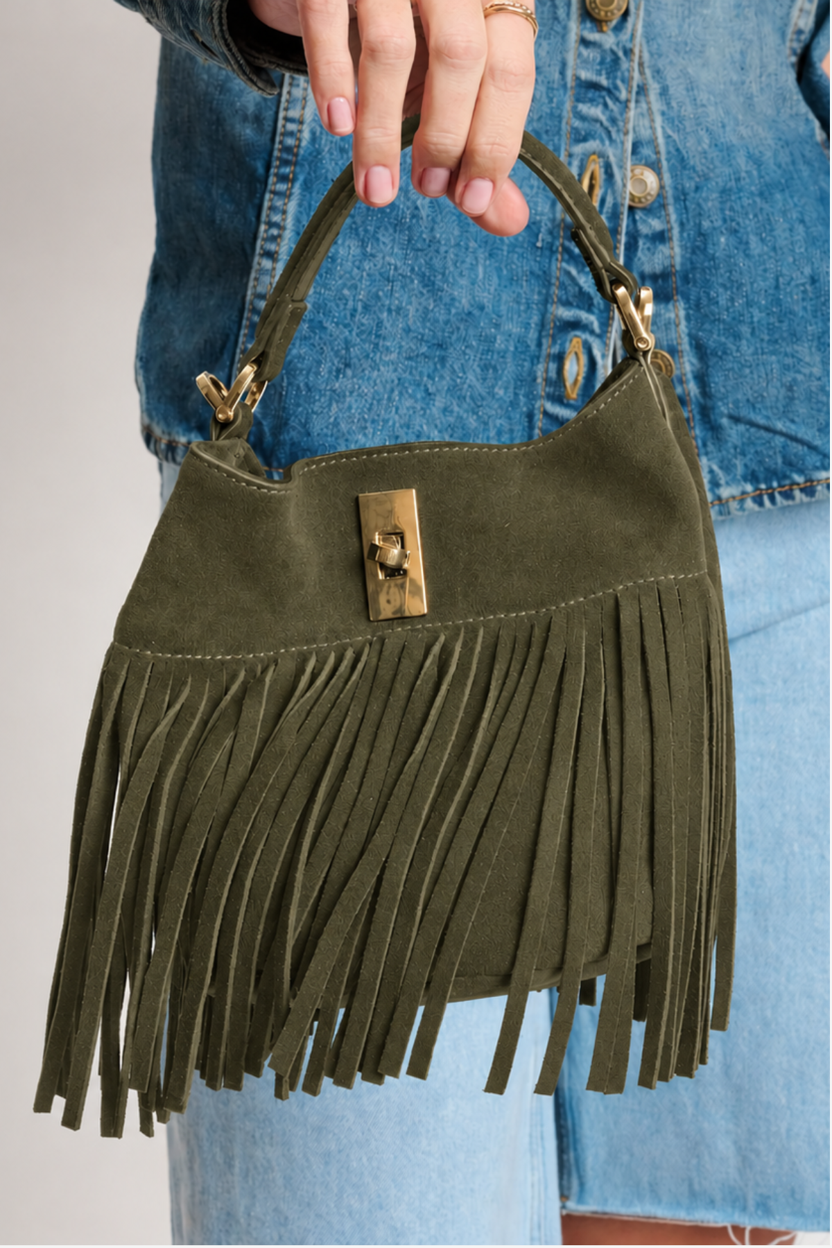 An olive green suede fringe handbag with gold-tone hardware, held by a person wearing a blue denim jacket and jeans.