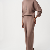 A model wearing a matching taupe-brown two-piece lounge set consisting of a mock-neck long-sleeve top and wide-leg trousers, paired with gray slip-on shoes, against a plain white studio background.