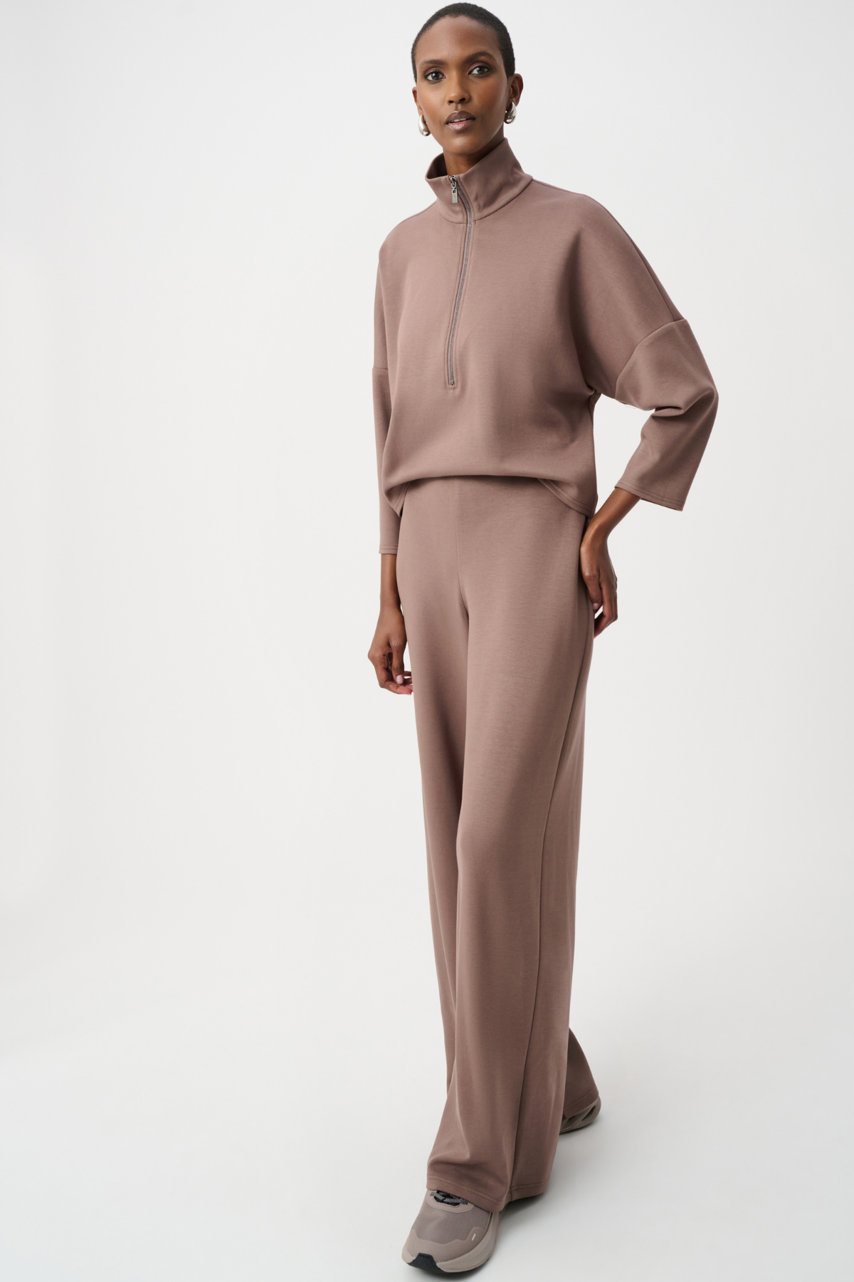 A model wearing a matching taupe-brown two-piece lounge set consisting of a mock-neck long-sleeve top and wide-leg trousers, paired with gray slip-on shoes, against a plain white studio background.