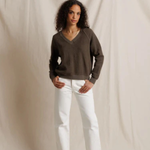 Woman wearing a brown sweater and white pants against a plain background