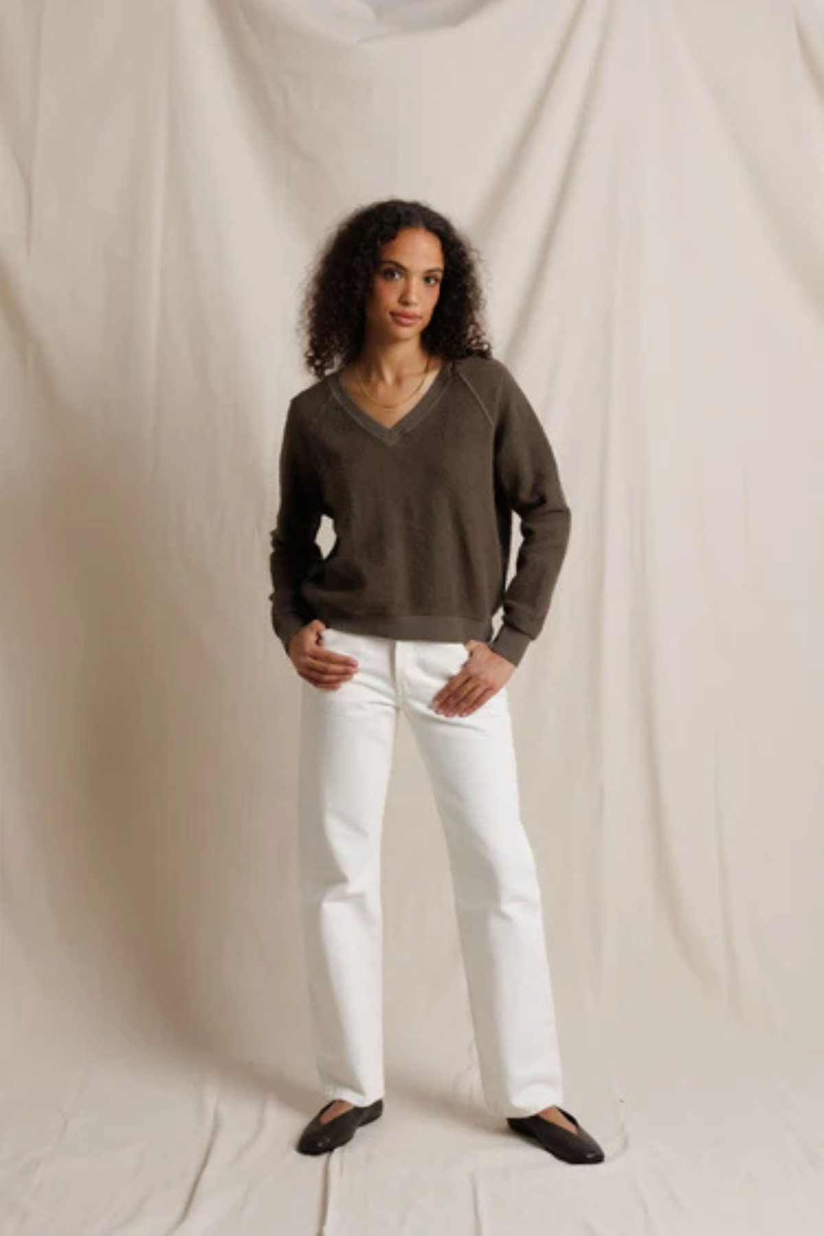 Woman wearing a brown sweater and white pants against a plain background