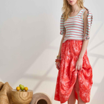 Woman wearing a striped shirt and red patterned skirt standing in a minimalistic room with wooden furniture.