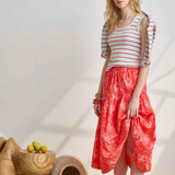 Woman wearing a striped shirt and red patterned skirt standing in a minimalistic room with wooden furniture.