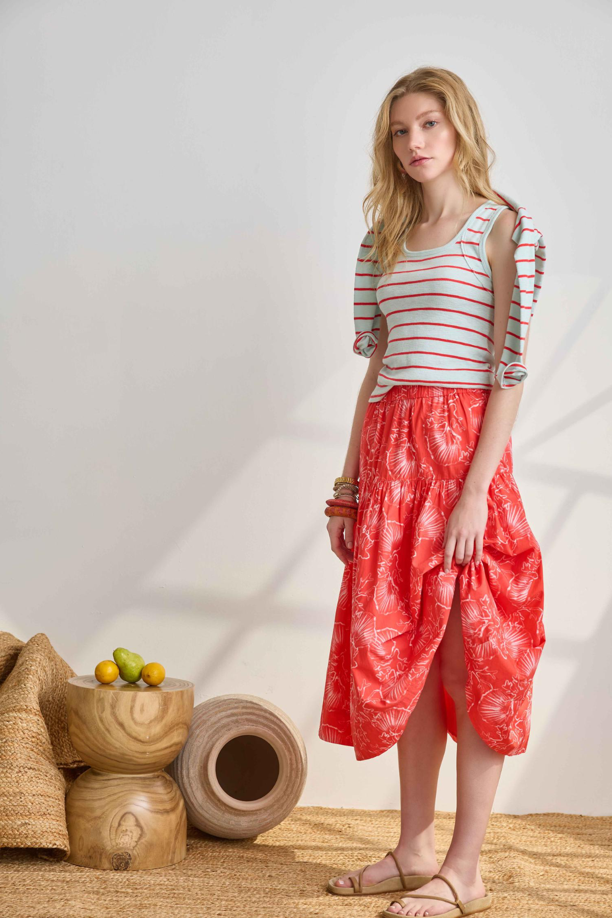 Woman wearing a striped shirt and red patterned skirt standing in a minimalistic room with wooden furniture.