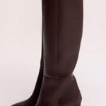 Dark brown knee-high leather boots with pointed toes and mid-height stiletto heels, shown on a model against a light beige studio background.