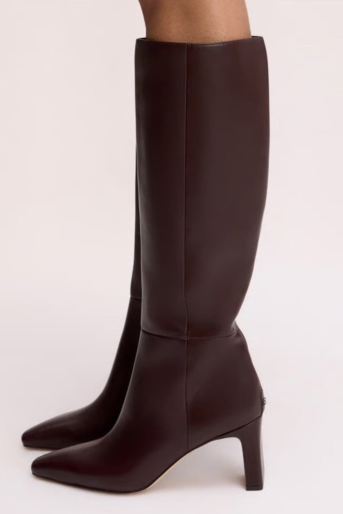 Dark brown knee-high leather boots with pointed toes and mid-height stiletto heels, shown on a model against a light beige studio background.