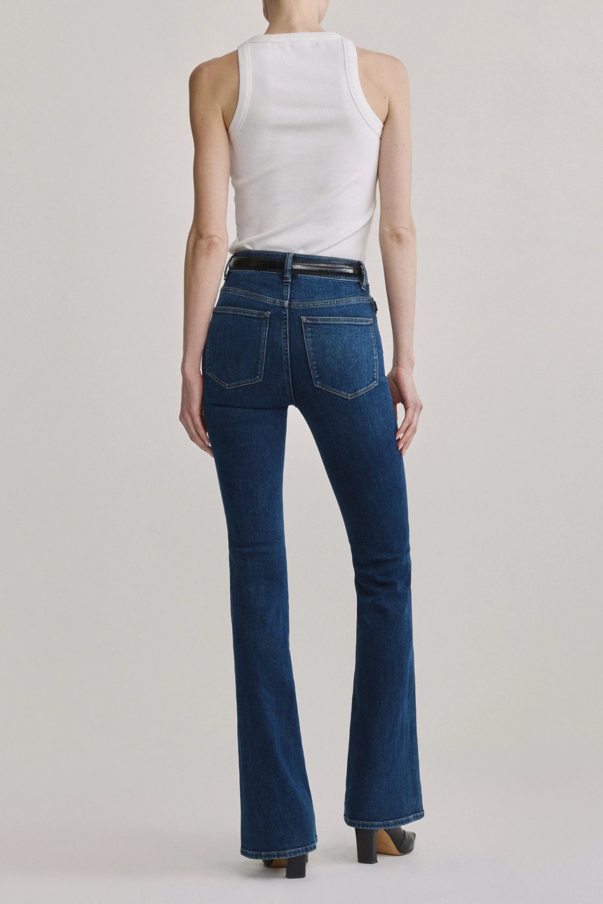 A model shown from the back wearing dark blue high-waisted flare jeans, a white sleeveless top, and brown heels against a light gray studio background.
