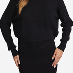 A woman wearing a black ribbed knit sweater and matching black ribbed knit skirt against a light gray studio background.