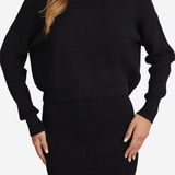 A woman wearing a black ribbed knit sweater and matching black ribbed knit skirt against a light gray studio background.
