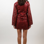 A woman viewed from behind wearing a short, shiny red mini dress with a textured snakeskin pattern and long sleeves, paired with brown ankle boots, against a plain light gray studio background.