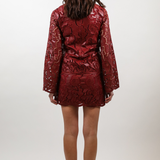 A woman viewed from behind wearing a short, shiny red mini dress with a textured snakeskin pattern and long sleeves, paired with brown ankle boots, against a plain light gray studio background.