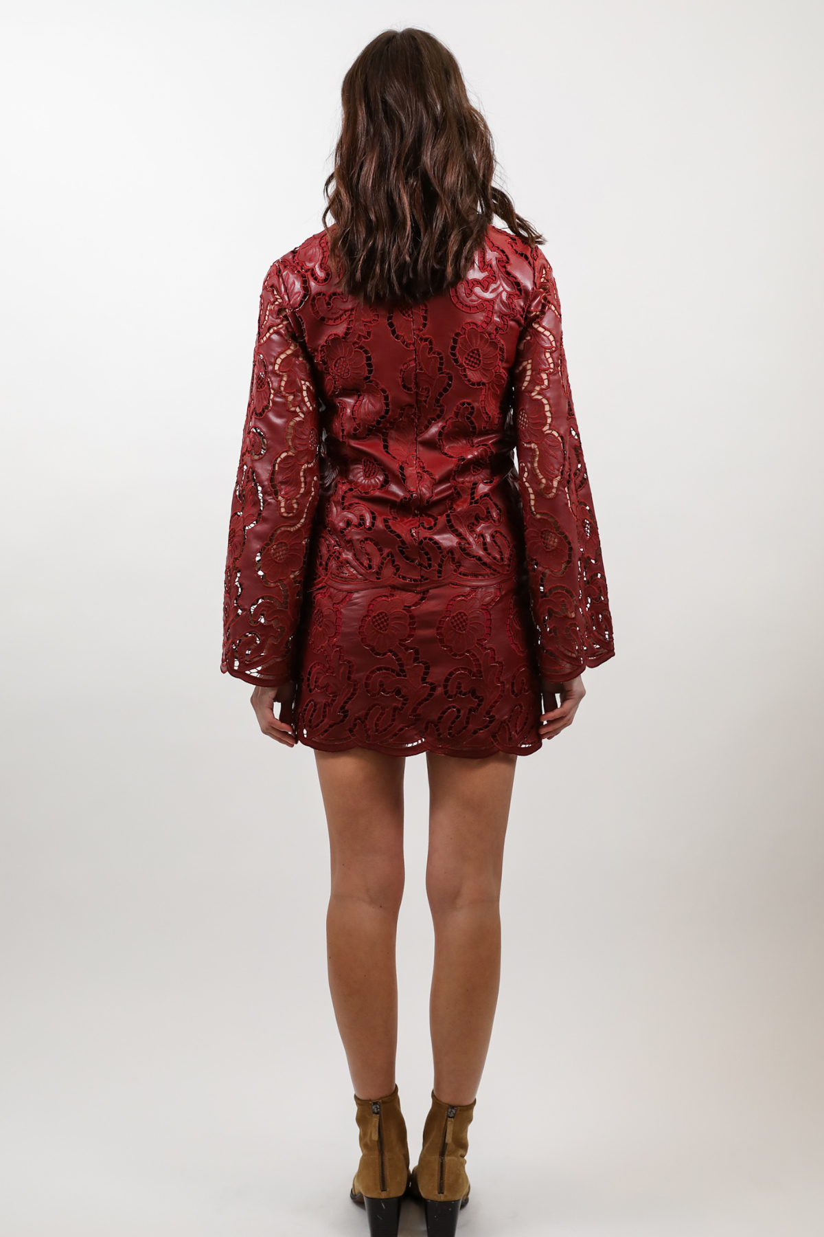 A woman viewed from behind wearing a short, shiny red mini dress with a textured snakeskin pattern and long sleeves, paired with brown ankle boots, against a plain light gray studio background.